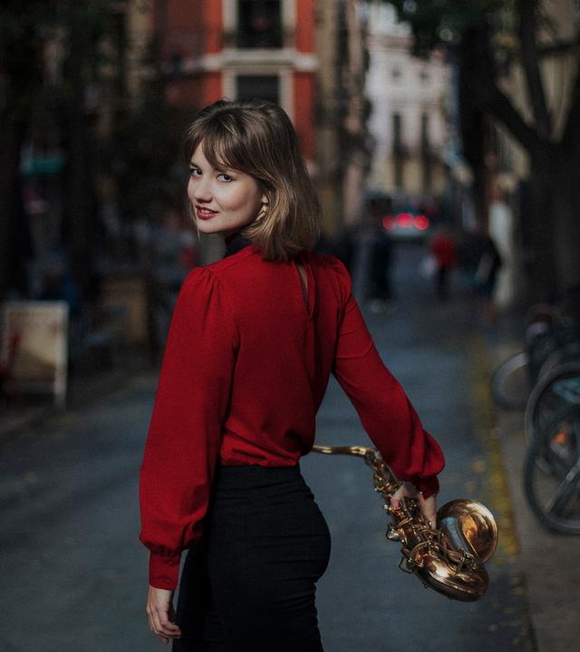 Die junge Saxophonistin Fabia Mantwill in roter Bluse und schwarzer Hose, von der Kamera auf der Straße in einer Wohngegend weglaufend, sich umdrehend in der Bewegung und das Saxophon in der rechten Hand habend.