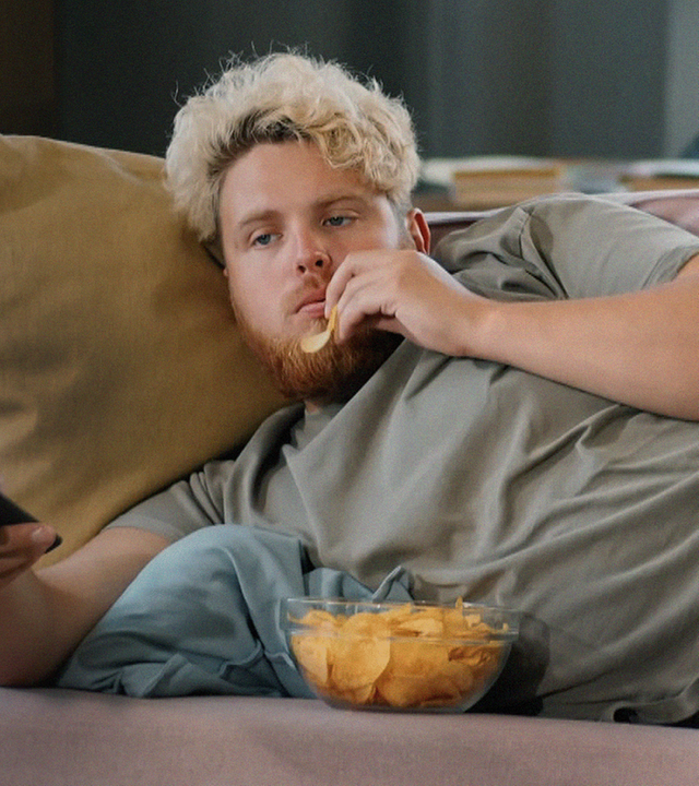 Junger Mann (Gen Z) liegt auf der Couch und isst Chips
