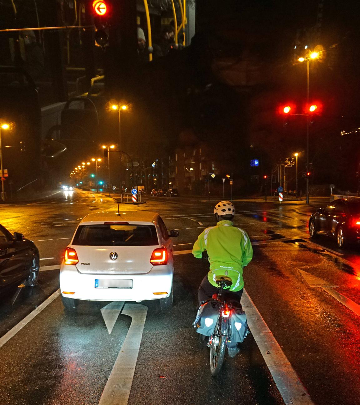 Fahrradfahrer mit Warnweste und Reflektoren wartet im Dunkeln an einer Ampel im Straßenverkehr.