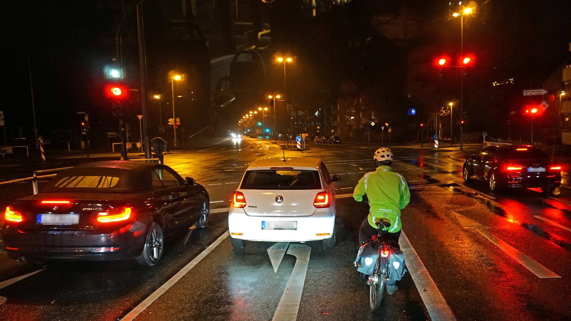 Fahrradfahrer mit Warnweste und Reflektoren wartet im Dunkeln an einer Ampel im Straßenverkehr.
