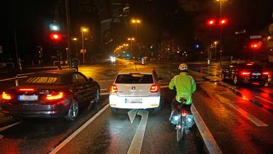 Fahrradfahrer mit Warnweste und Reflektoren wartet im Dunkeln an einer Ampel im Straßenverkehr.
