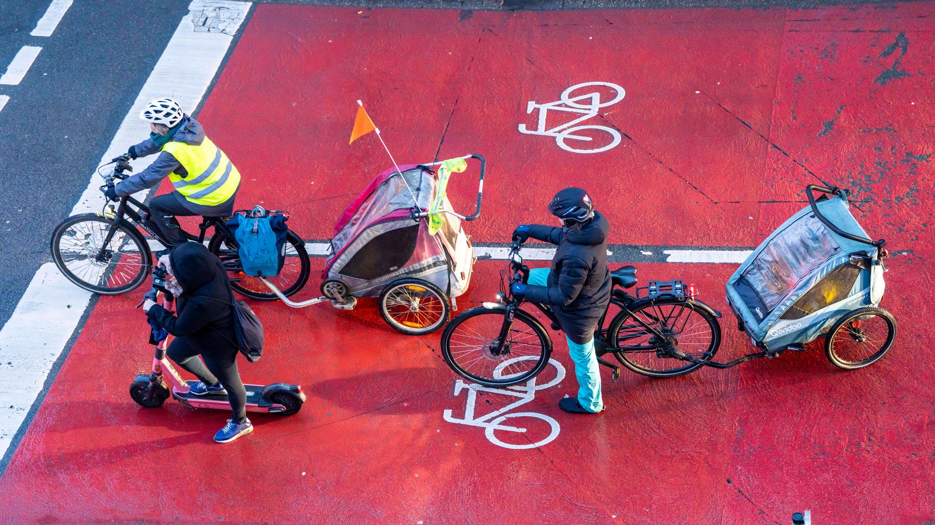 Ein Scooter-Fahrer und zwei Radfahrer mit Fahrradanhängern warten an einer Kreuzung, von oben fotografiert.