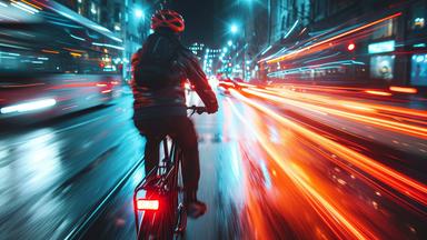 Ein Fahrradfahrer im Dunkeln auf einer Straße, links und rechts neben ihm durch Langzeitbelichtung hervorgerufene Lichtstreifen-Effekte von Auto-Rücklichtern.