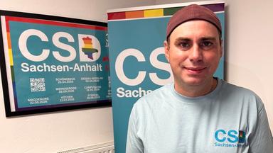 Falko Jentsch steht vor einem Schild mit der Aufschrift "CSD Sachsen-Anhalt".