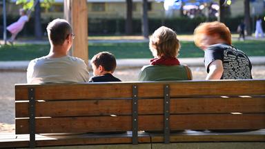 Eine Familie sitzt auf einer Holzbank an einem Spielplatz im Park. (Symbolbild)