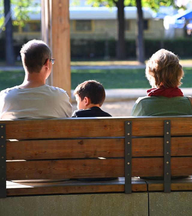Eine Familie sitzt auf einer Holzbank an einem Spielplatz im Park. (Symbolbild)