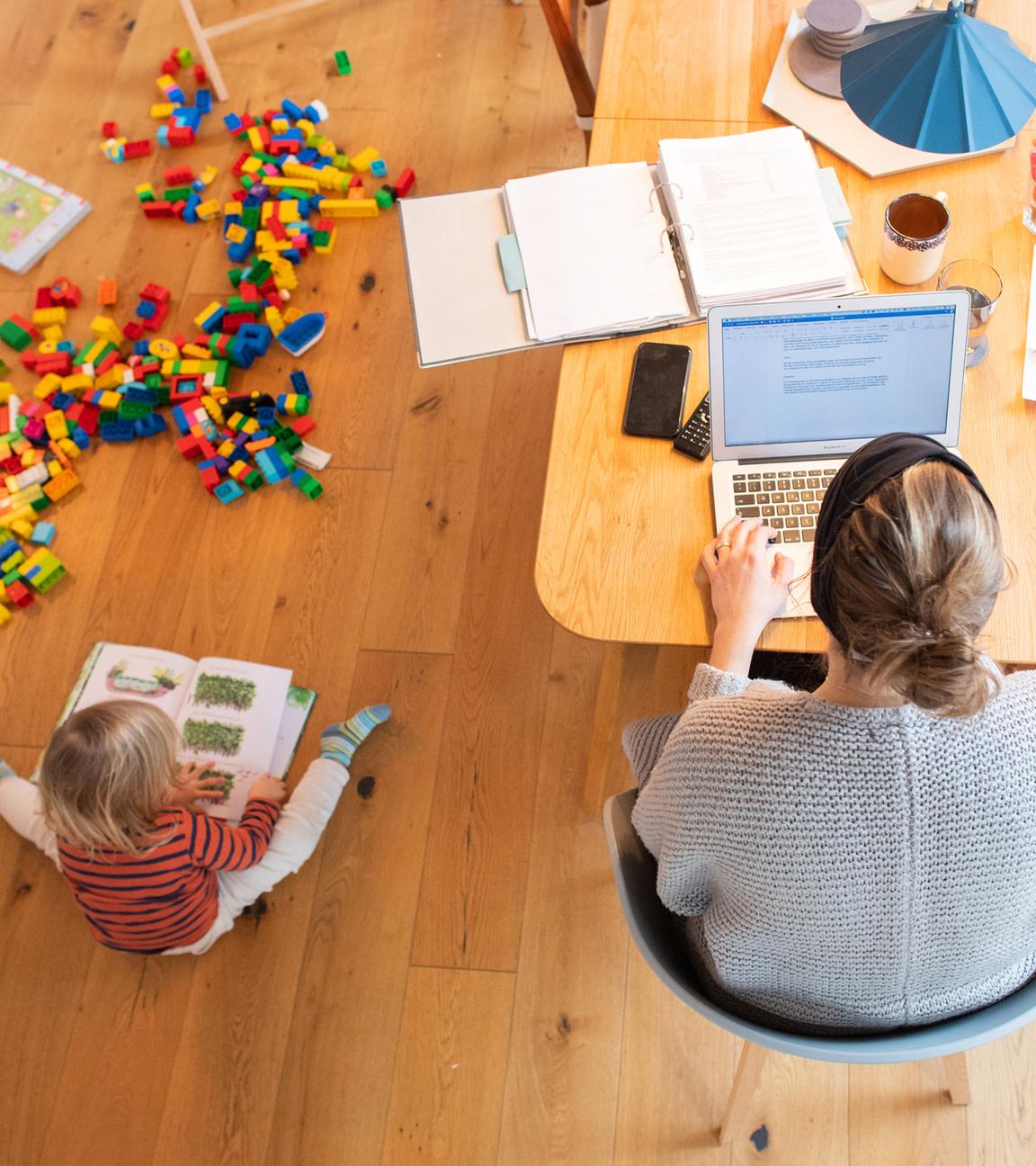 Ein zweijähriges Kind spielt im Wohnzimmer, während seine Mutter Zuhause im Homeoffice an einem Laptop arbeitet.