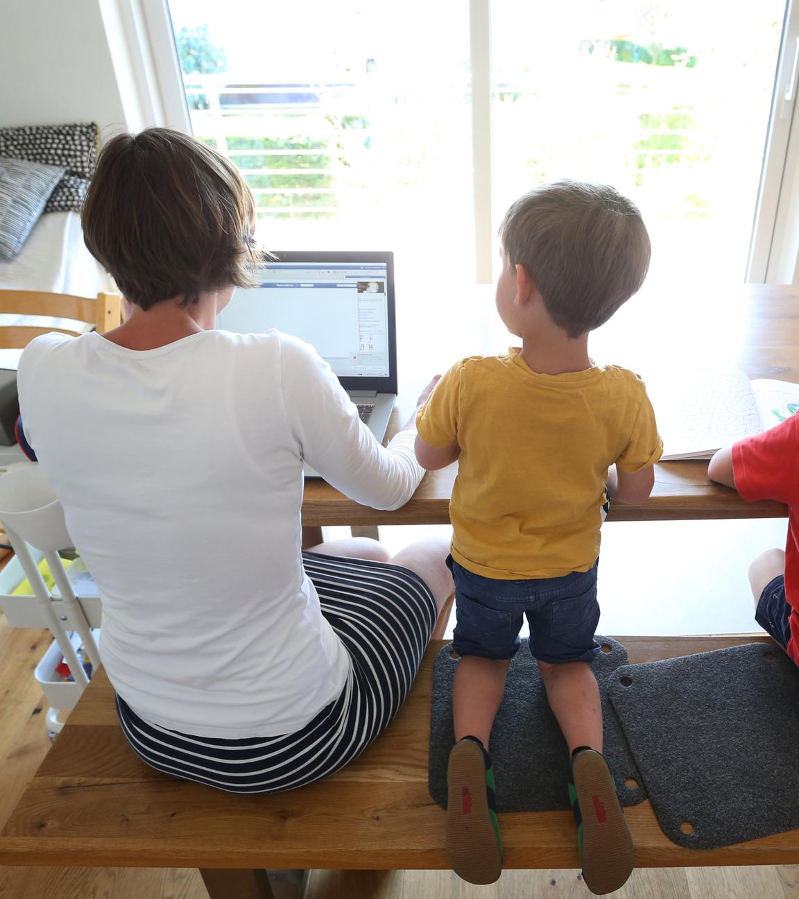 Die Mutter arbeitet Zuhause an einem Laptop, während ihre Kinder neben ihr malen und ein Buch ansehen.