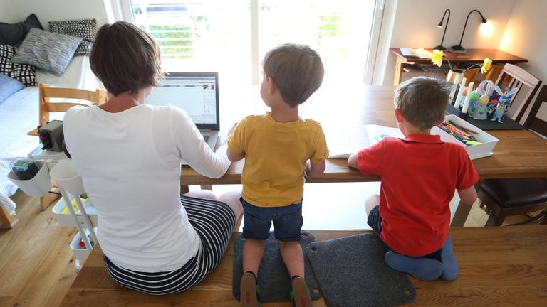Die Mutter arbeitet Zuhause an einem Laptop, während ihre Kinder neben ihr malen und ein Buch ansehen.