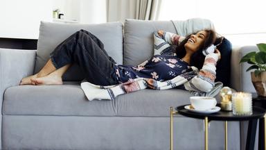 Eine Frau trägt weiße drahtlose Kopfhörer, hört Musik, ruht sich aus und liegt auf der Couch im Wohnzimmer