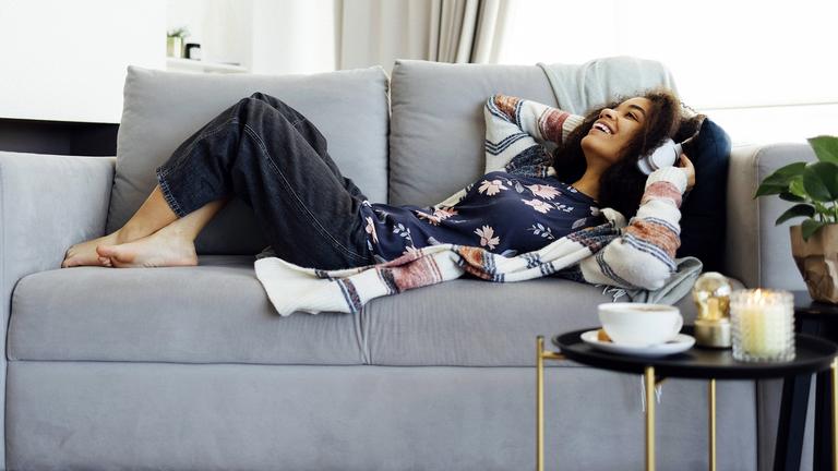 Eine Frau trägt weiße drahtlose Kopfhörer, hört Musik, ruht sich aus und liegt auf der Couch im Wohnzimmer
