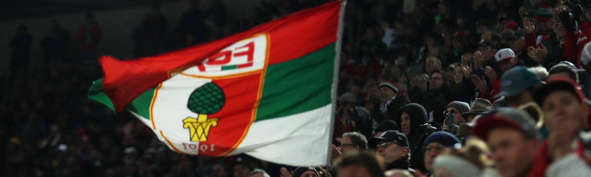 Es sind FC Augsburg Fans zu sehen, wie sie eine Flagge mit dem Vereinslogo hochhalten.