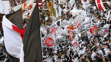 Auf einer Tribüne jubeln Fußballfans in braun, rot und weiß. Es wehen Fahnen weißes Konfetti.