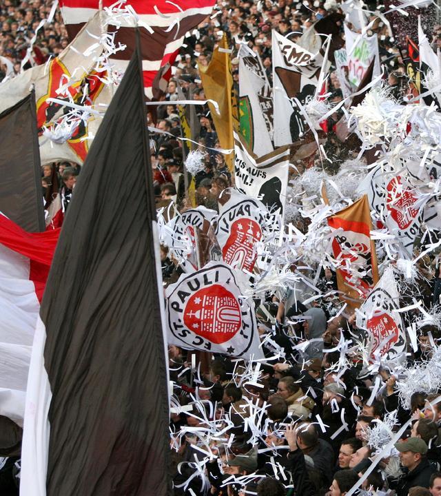 Auf einer Tribüne jubeln Fußballfans in braun, rot und weiß. Es wehen Fahnen weißes Konfetti.
