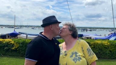 Das Ehepaar Baxter steht am Ufer und küsst sich auf die Lippen. Im Hintergrund liegen mehrere Boote im Meer.