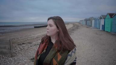 Ellena Baxter blickt an einem Strand mit Strandhütten nachdenklich aufs Meer.