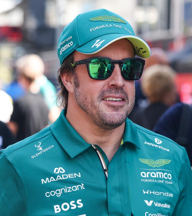 Fernando Alonso läuft mit Sonnenbrille, Kappe und grünem Aston Martin Shirt in Richtung Kamera