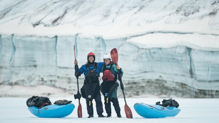Zwei Personen mit Paddeln in der Hand stehen zwischen Kanus in eisiger Landschaft.