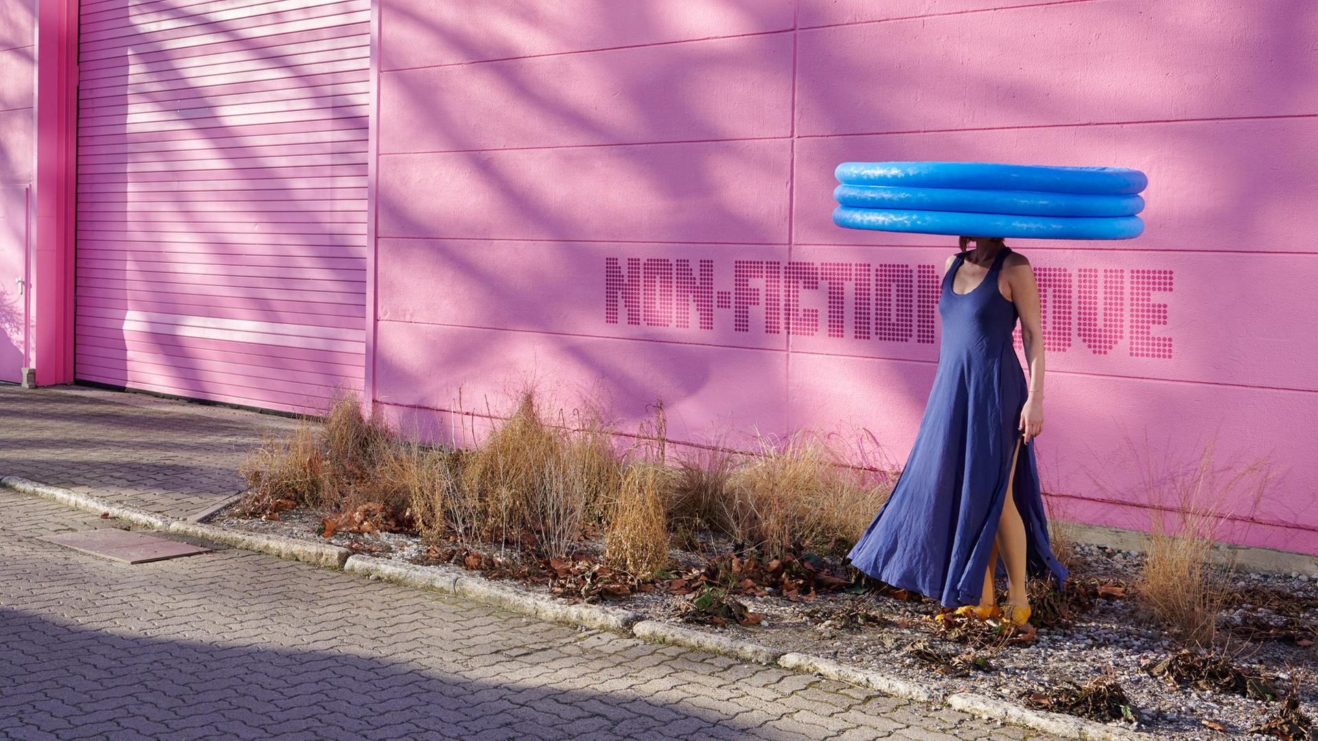 Vor einer pink-farbenen Wand steht eine Frau, die ein blaues Kleid trägt. Sie trägt ein aufblasbares Planschbecken auf dem Kopf.