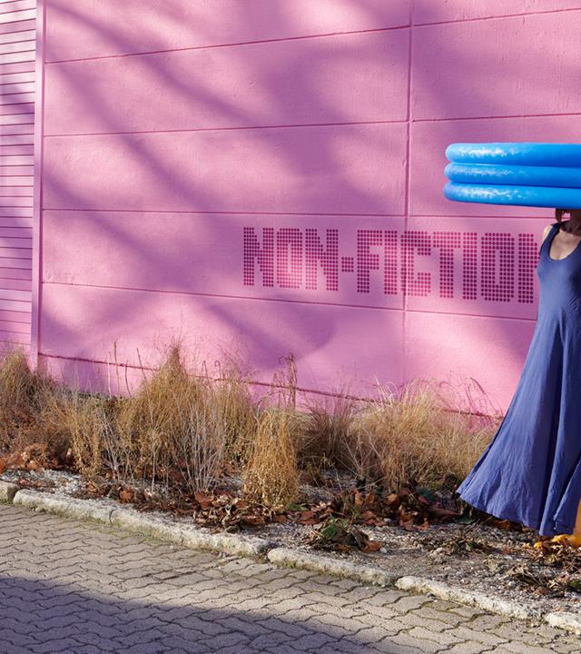 Vor einer pink-farbenen Wand steht eine Frau, die ein blaues Kleid trägt. Sie trägt ein aufblasbares Planschbecken auf dem Kopf.