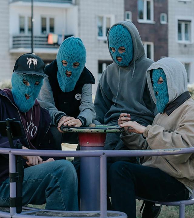 Vier junge Männer tragen blaue Strickmasken und sitzen auf einem Kinderkarussel.
