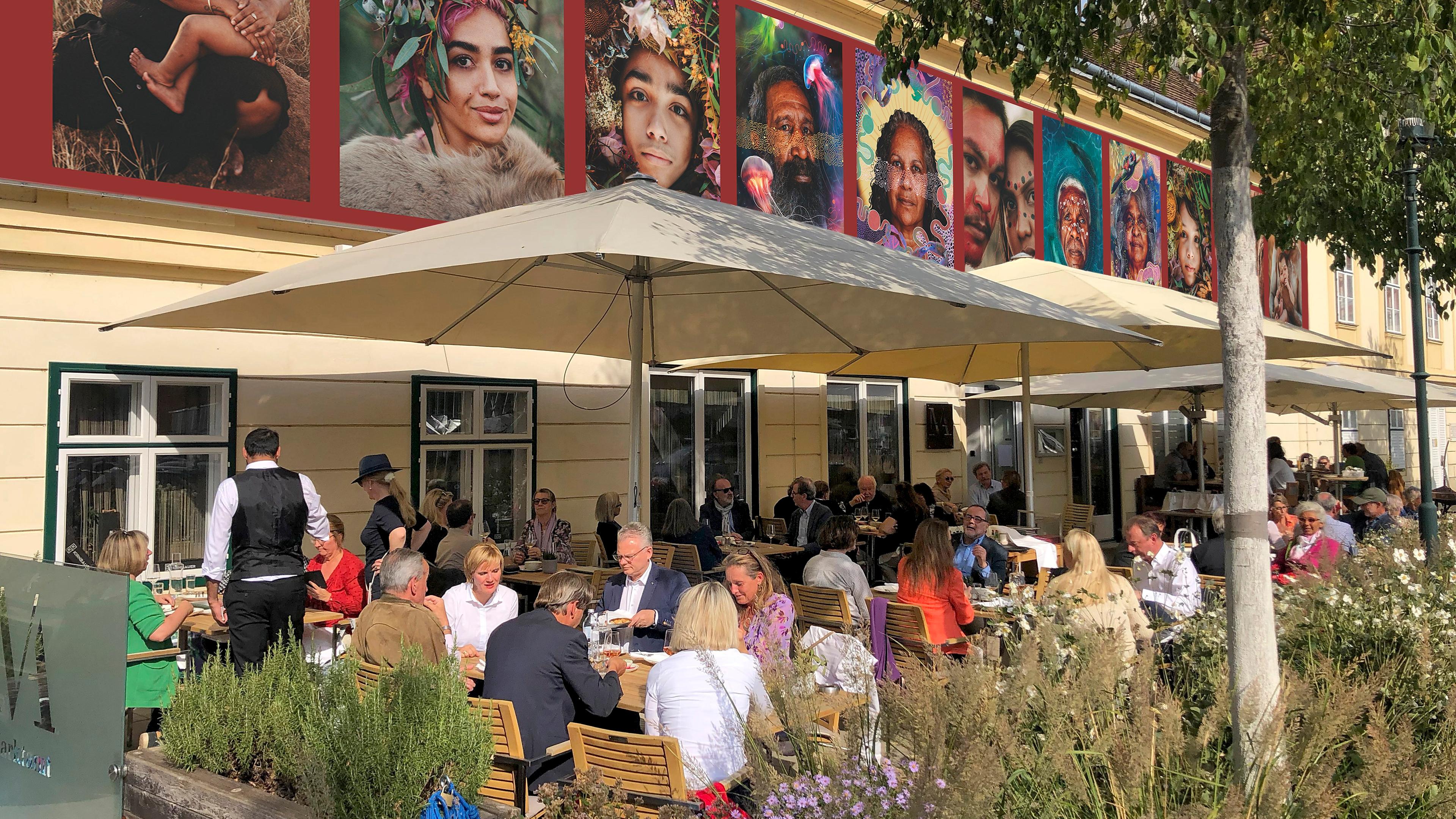 Man sieht ein belebtes Straßencafe. Auf Höhe des ersten Stockwerkes des Cafes sind großformatige Fotografien zu sehen.