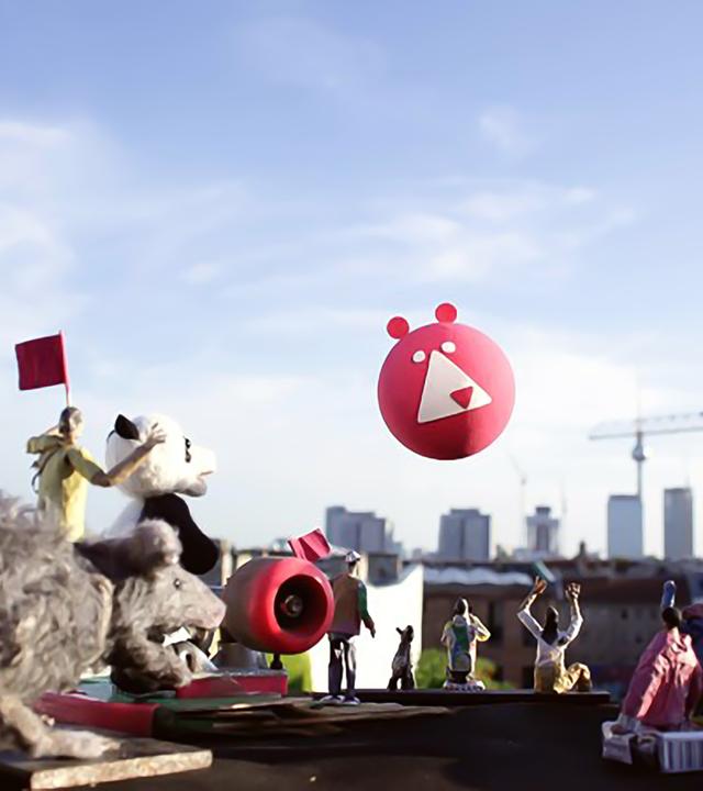 Viele kleine Figuren und Kinderspielzeuge stehen auf einem Tisch und blicken in Richtung einer Skyline, wo auch ein Ballon langfliegt.