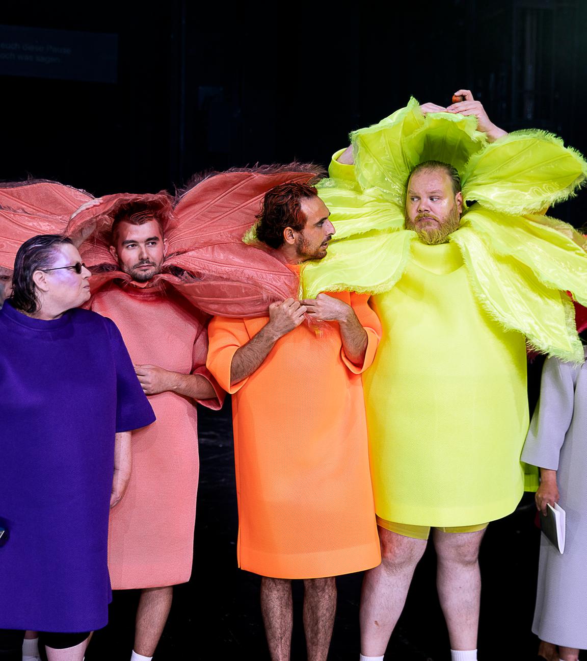 Bühnenszene: Menschen tragen bunte Blumenkostüme vor schwarzem Hintergrund.