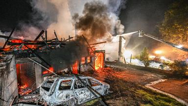 Auf dem Bild ist zu sehen: Vor den rauchenden Resten des Schwarzwaldhofs steht ein ausgebranntes Auto. Dahinter glühen Balken und Trümmer rot. In mehreren Öffnungen sind Flammen zu sehen.