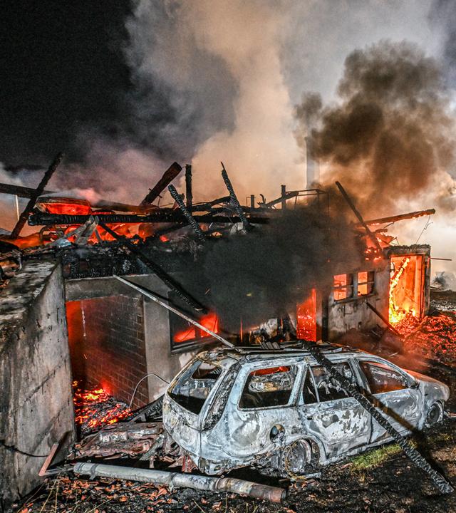 Auf dem Bild ist zu sehen: Vor den rauchenden Resten des Schwarzwaldhofs steht ein ausgebranntes Auto. Dahinter glühen Balken und Trümmer rot. In mehreren Öffnungen sind Flammen zu sehen.