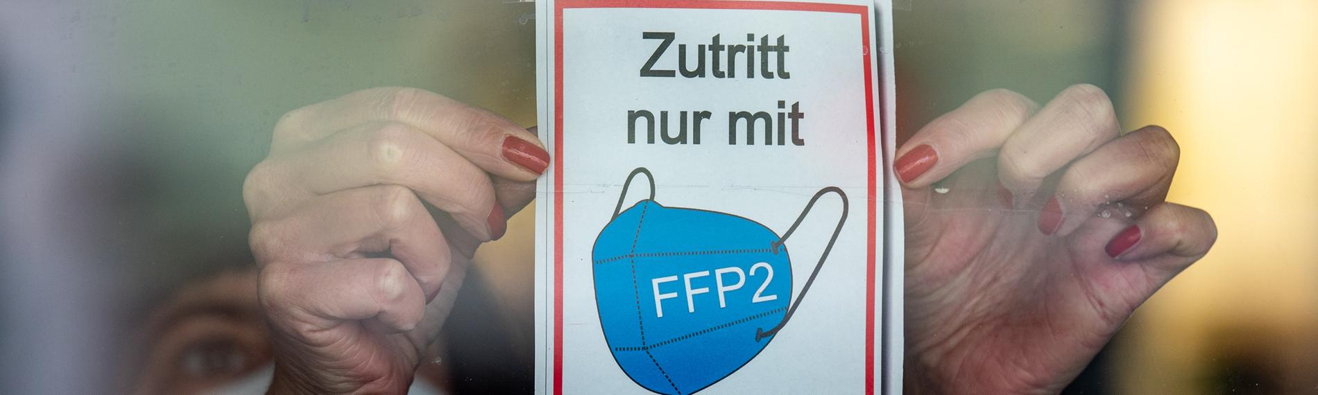  Bayern, Straubing: Eine Frau klebt ein Hinweisschild mit der Aufschrift ·Zutritt nur mit FFP2 Maske· an eine Glastür am Eingang eines Geschäfts in der Innenstadt.
