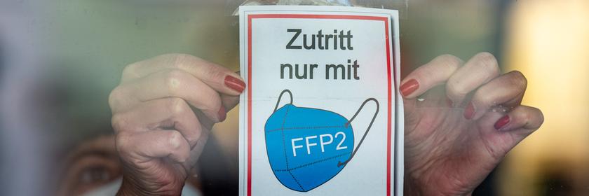  Bayern, Straubing: Eine Frau klebt ein Hinweisschild mit der Aufschrift ·Zutritt nur mit FFP2 Maske· an eine Glastür am Eingang eines Geschäfts in der Innenstadt.