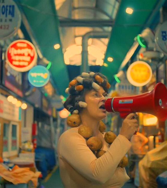 Eine Frau mit Lockenwicklern im Haar spricht durch ein Megafon, während sie durch einen koreanischen Markt geht.