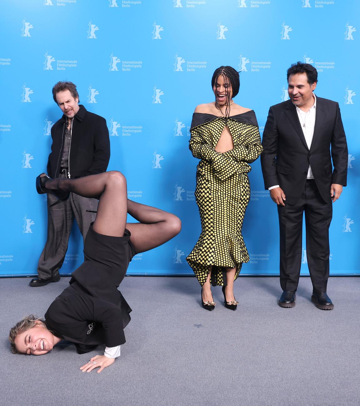Vor dem blauen Hintergrund der Berlinale-Pressekonferenzen stehen mehrere Personen und schauen lachend zu einer Frau im schwarzen Anzug mit kurzen Hosen und Stöckelschuhen, die auf dem Boden Break-Dance vorführt.