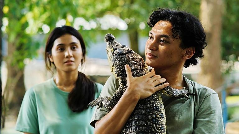 Ein junger  Mann trägt ein kleines Krokodil auf dem Arm, links hinter ihm steht eine junge Frau.