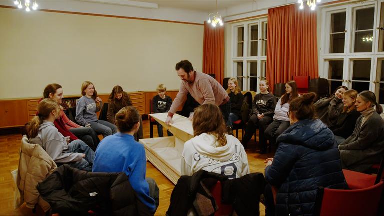 Eine Schulergruppe sitzt in einem Stuhlkreis um einen Sarg, von dem ein Mann den Deckel abnimmt.