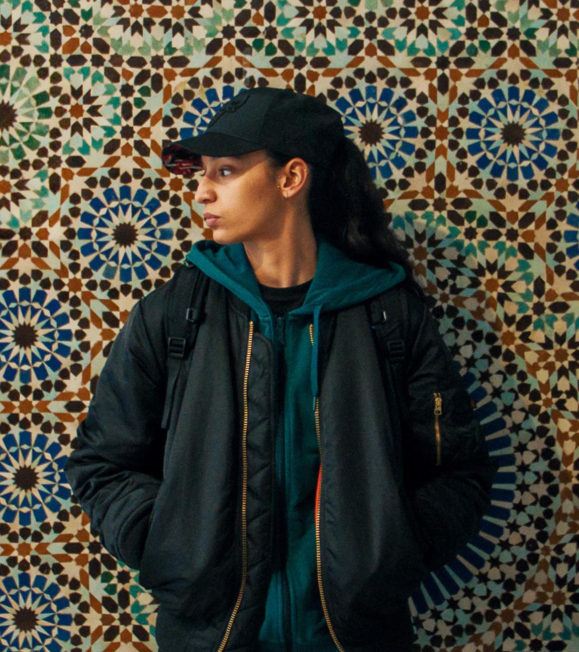 Eine junge Frau lehnt an einer bunten, Mosaik-Wand. Sie trägt eine schwarze Kappe, die dunklen Haare in einem Zopf und ein dunkles Outfit. Sie blickt zur Seite.