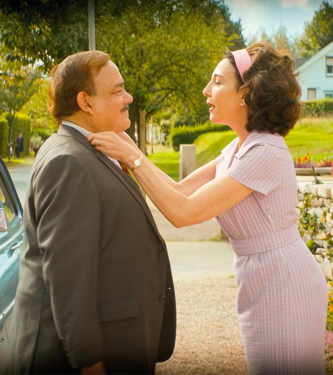 Ein Mann und eine Frau stehen einander gegenüber und sind für den Betrachter deswegen nur von der Seite zu sehen. Der Mann trägt einen Anzug und die Frau ein pastellfarbenes lila Kleid mit passendem Haarband. Die Frau richtet den Hemdkragen des Mannes, während sie mit ihm redet.