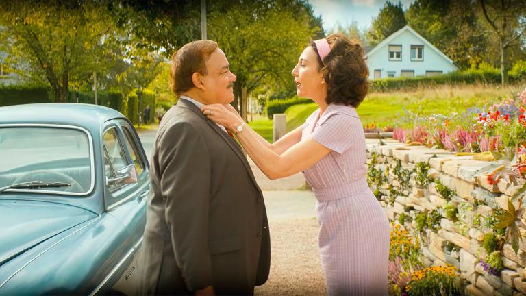 Ein Mann und eine Frau stehen einander gegenüber und sind für den Betrachter deswegen nur von der Seite zu sehen. Der Mann trägt einen Anzug und die Frau ein pastellfarbenes lila Kleid mit passendem Haarband. Die Frau richtet den Hemdkragen des Mannes, während sie mit ihm redet.