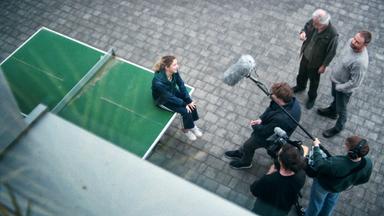 Ein junges Mädchen sitzt auf einer Tischtennisplatte. Vor ihr stehen fünf Männer mit Kamera und Mikrofonen, die sie zu interviewen scheinen.