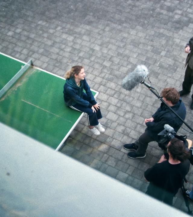 Ein junges Mädchen sitzt auf einer Tischtennisplatte. Vor ihr stehen fünf Männer mit Kamera und Mikrofonen, die sie zu interviewen scheinen.