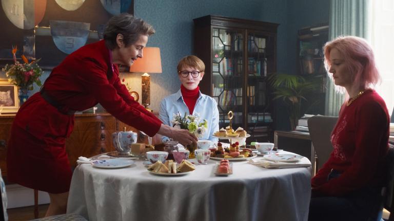 Zwei Frauen sitzen steif an einem gedeckten Kaffe-Tisch. Eine dritte steht daneben und stellt eine kleine Blumenvase in die Mitte.