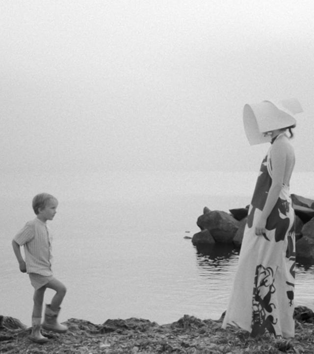 Auf einer Schwarz-Weiß-Fotografie sieht man drei Kinder und zwei Foto-Models in futuristischer Bekleidung am Strand.