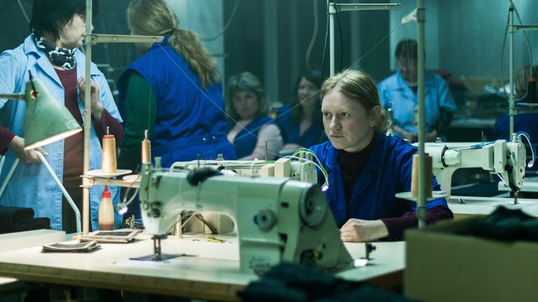 Eine blonde Frau in einem blauen Kittel sitzt in einer Werkshalle an einer Nähmaschine und schaut nachdenklich in die Ferne.