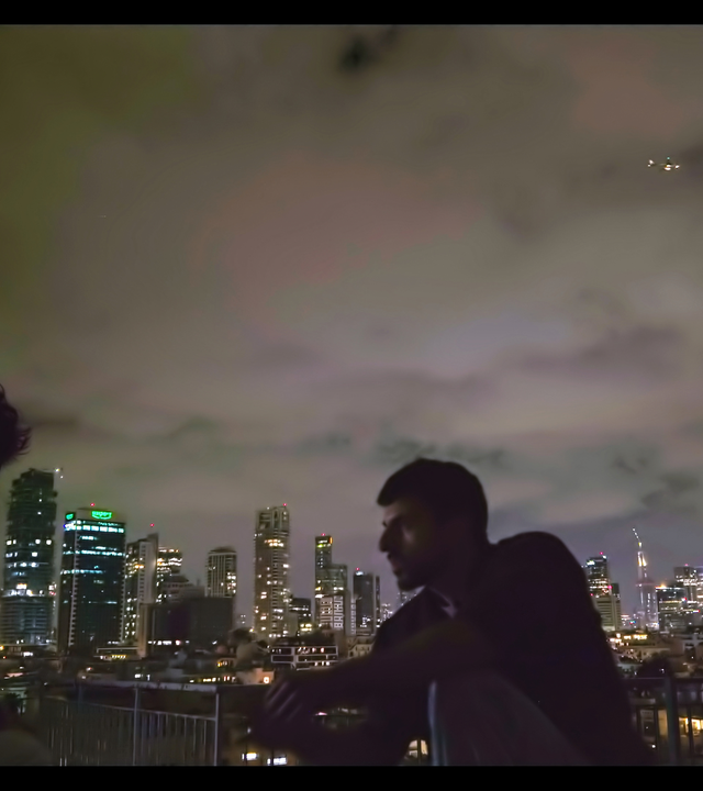 Zwei Männer sitzen abends vor der Skyline von Tel Aviv mit Hochhäusern. Sie sind nur als dunkle Silhouetten zu sehen.