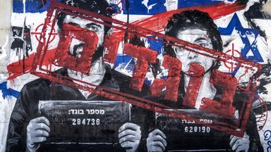 Auf einem Mural sind die Porträts eines Mannes und einer Frau zu sehen, die wie bei Verbrecherfotos Schilder mit Nummern tragen. Die Gesichter wurden mit hebräischer Schrift übermalt.