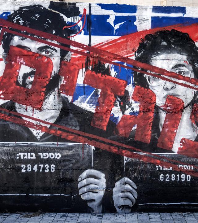 Auf einem Mural sind die Porträts eines Mannes und einer Frau zu sehen, die wie bei Verbrecherfotos Schilder mit Nummern tragen. Die Gesichter wurden mit hebräischer Schrift übermalt.