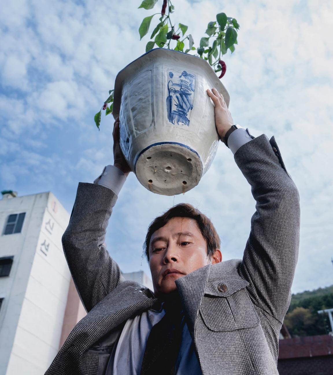 Ein Mann mittleren Alters in einer Tweed-Jacke hebt einen großen Blumentopf über den Kopf, als ob er ihn gleich werfen möchte.