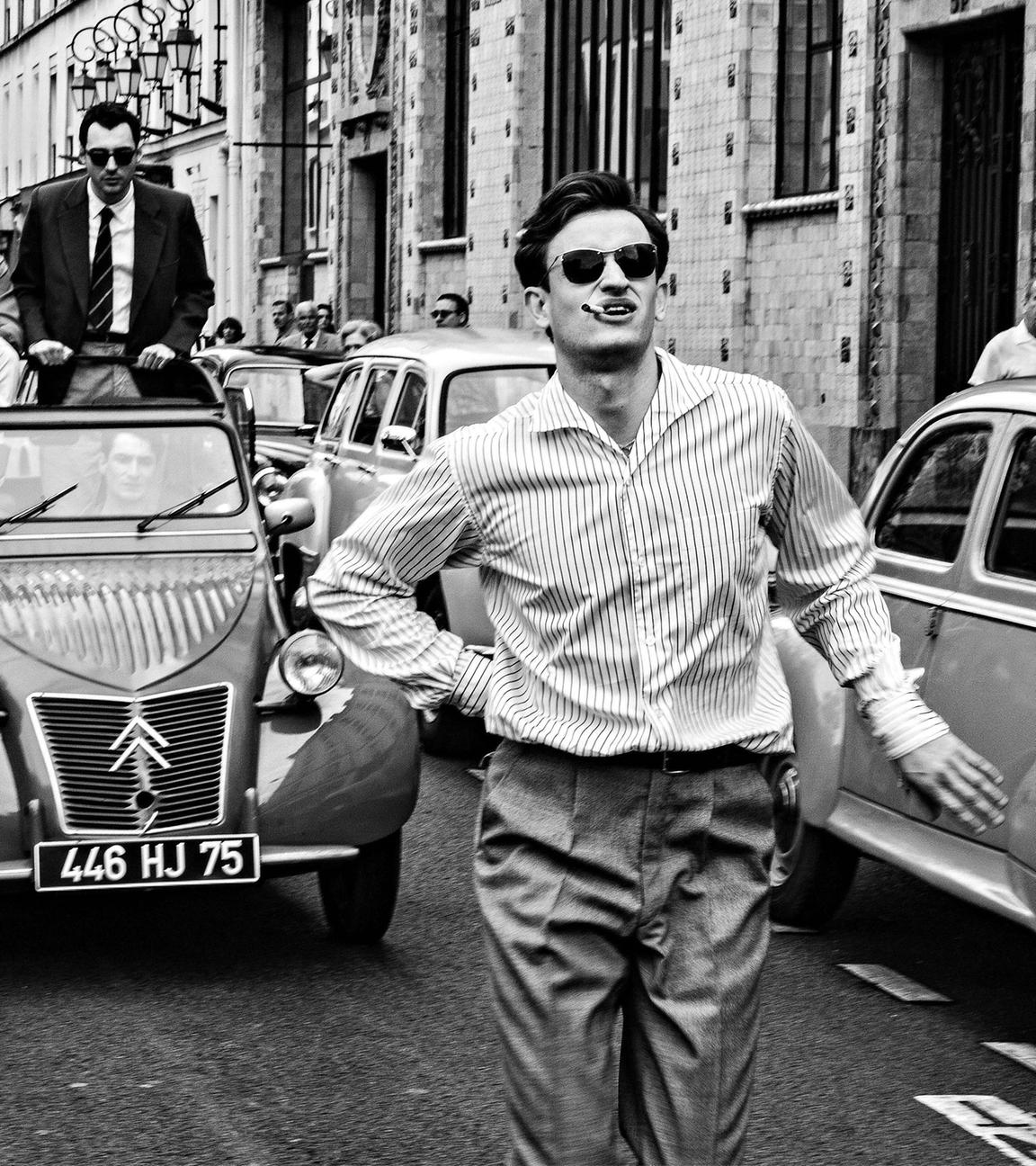 Das Schwarz-Weiß-Bild zeigt einen jungen Mann in einem Hemd, der mit Zigarette im Mund und Sonnenbrille zwischen Autos aus den 1950er Jahren auf einer Straße läuft. Hinter ihm auf der Straße fährt eine Cabrio-Ente mit offenem Dach. Drei Leute stehen in dem Auto, eine Person hat eine Kamera und filmt den Mann auf der Straße von hinten.
