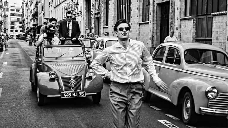 Das Schwarz-Weiß-Bild zeigt einen jungen Mann in einem Hemd, der mit Zigarette im Mund und Sonnenbrille zwischen Autos aus den 1950er Jahren auf einer Straße läuft. Hinter ihm auf der Straße fährt eine Cabrio-Ente mit offenem Dach. Drei Leute stehen in dem Auto, eine Person hat eine Kamera und filmt den Mann auf der Straße von hinten.